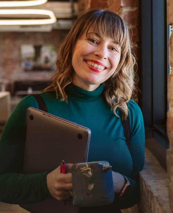 一位女士站在窗户旁边，面带微笑，手里拿着一台关闭的笔记本电脑和一个咖啡杯
