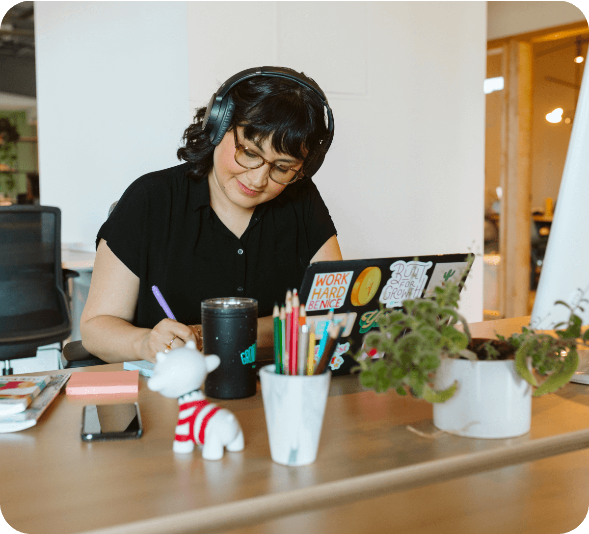 woman with headphones and glasses sitting at a desk takes notes in front of her laptop