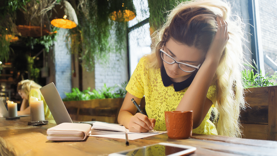 a woman wearing glasses writes notes at a desk in a sunny cafe