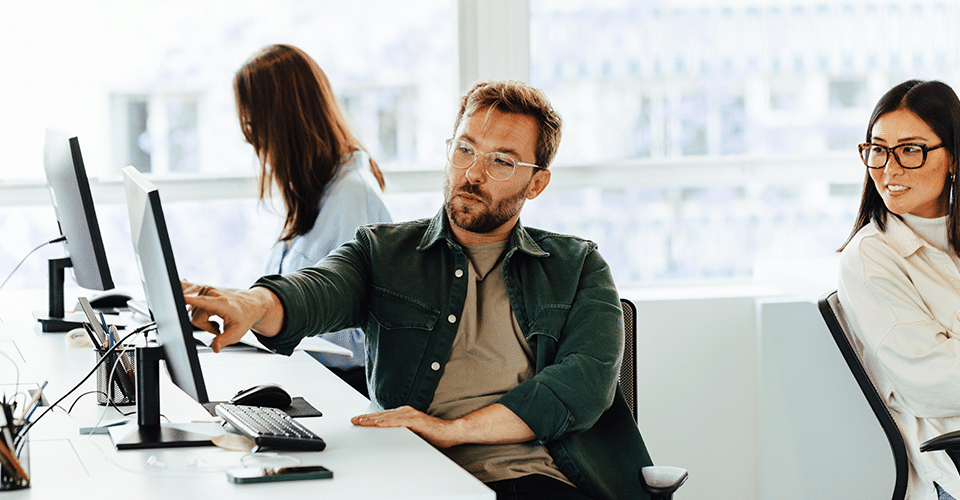 a man points to his computer screen to ask the opinion of a female coworker in a brightly lit office space