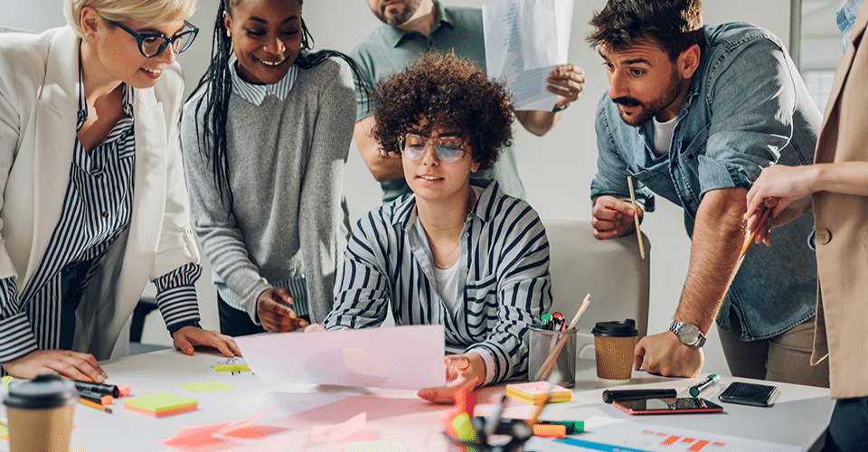 a group of creatives huddle around a woman holding a piece of paper as they all discuss a design