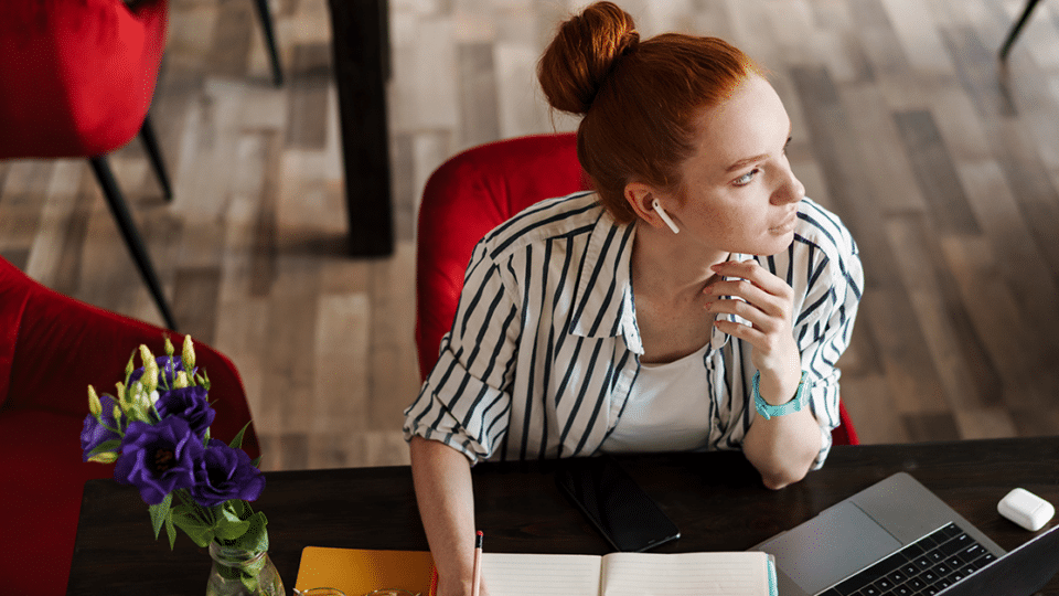 a red-haired woman writes notes in a notepad while working at her desk
