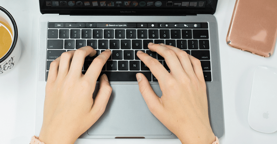 hands type on a laptop keyboard next to a cup of coffee and a wireless mouse
