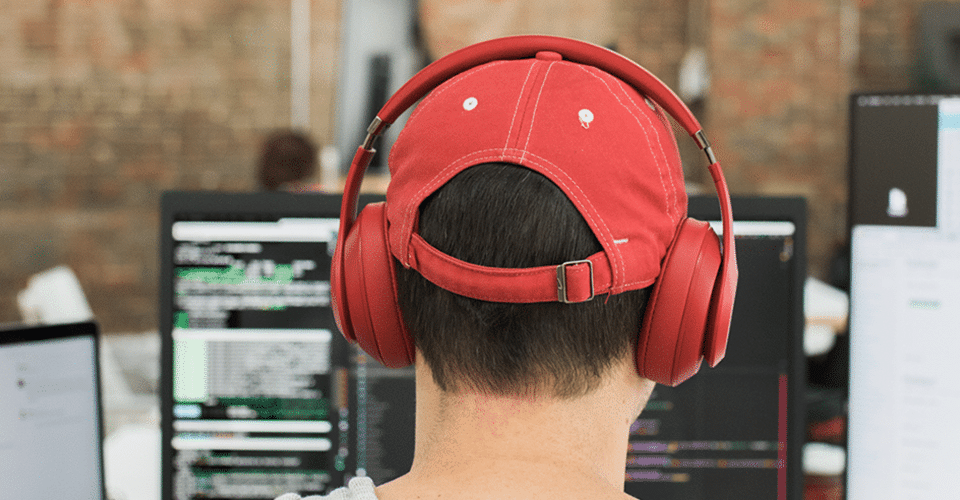 photo of the back of a man's head. He wears a red baseball cap and red over-the-ear headphones while he works on code
