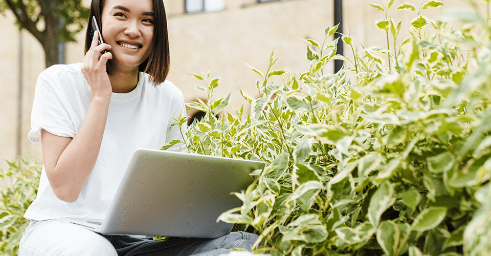 a woman works outside next to a bunch of greenery. she talks on the phone while holding a laptop