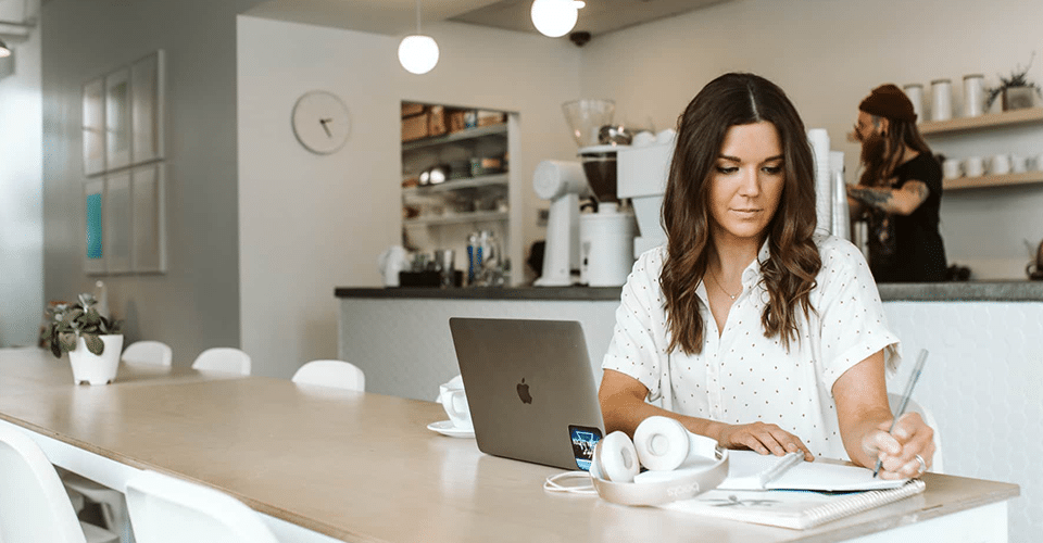 A woman uses a free invoice generator at a coffee shop, while a man at the counter behind her works on creating a free invoice template.
