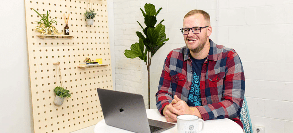 a man in a red and blue flannel shirt sits in front of his laptop at a table in a white office space