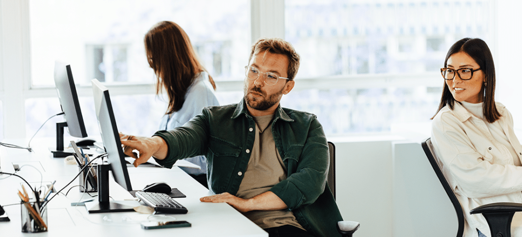a man points to his computer screen to ask the opinion of a female coworker in a brightly lit office space