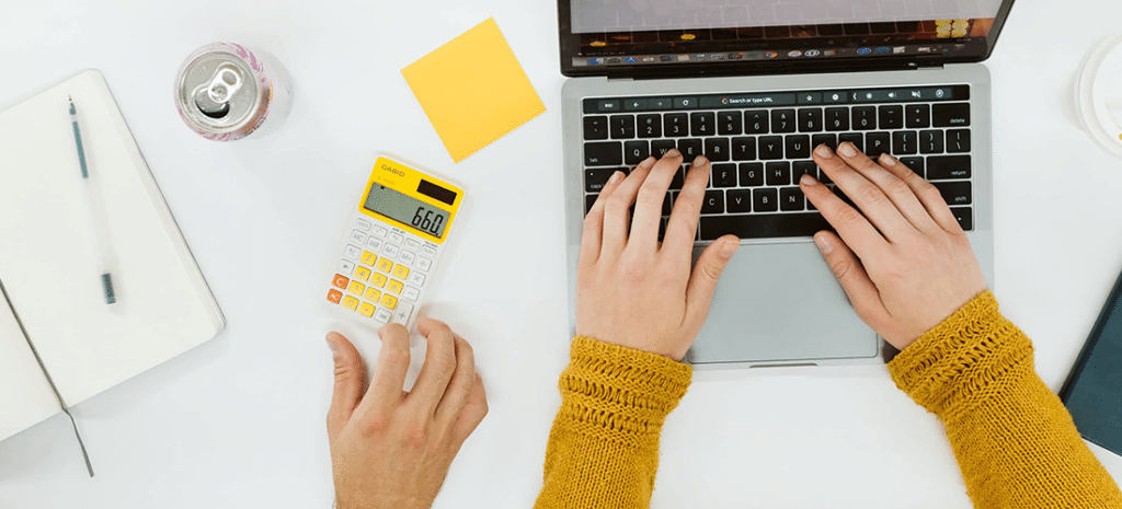 overhead shot of arms wearing yellow sleeves are poised above a laptop keyboard, ready to type. to the left of the computer, a calculator, post it notes, a notebook, and another person's hand are visible