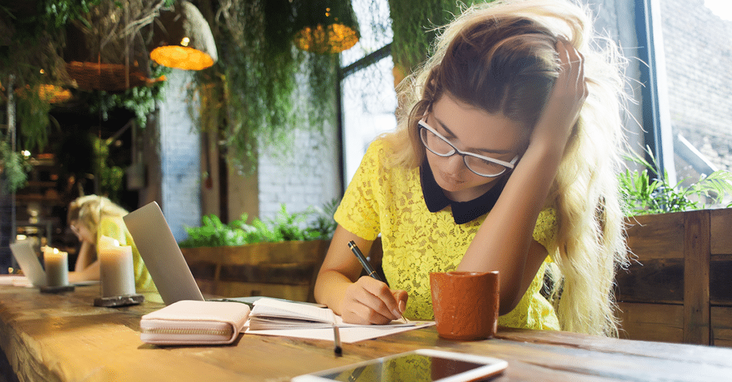 a woman wearing glasses writes notes at a desk in a sunny cafe
