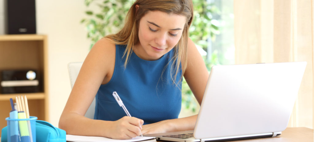 Photograph of girl writing in notebook while learning on computer