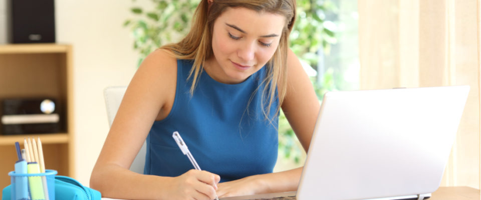 Photograph of girl writing in notebook while learning on computer