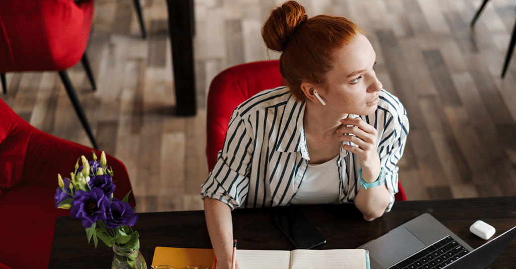 a red-haired woman writes notes in a notepad while working at her desk