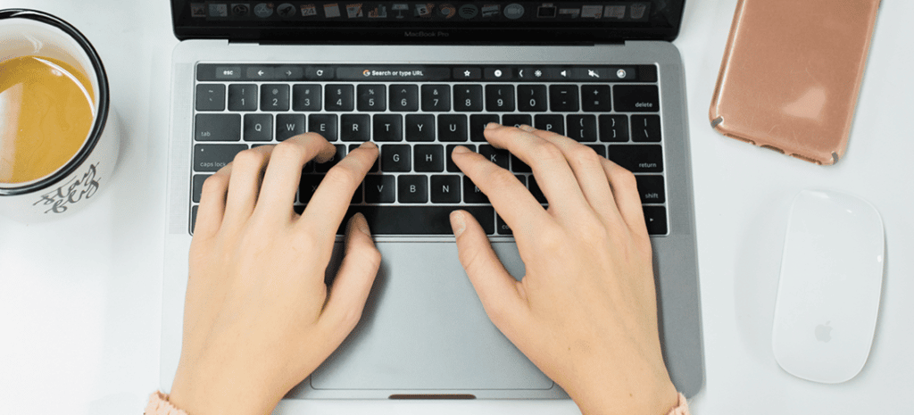 hands type on a laptop keyboard next to a cup of coffee and a wireless mouse