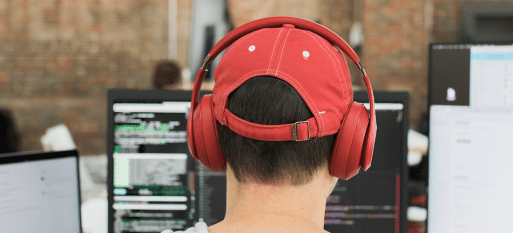 photo of the back of a man's head. He wears a red baseball cap and red over-the-ear headphones while he works on code
