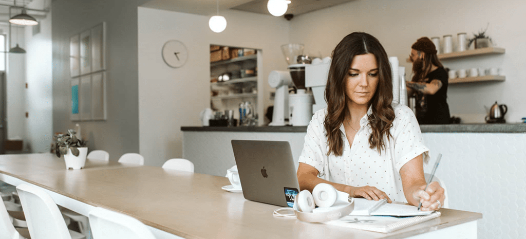 A woman uses a free invoice generator at a coffee shop, while a man at the counter behind her works on creating a free invoice template.