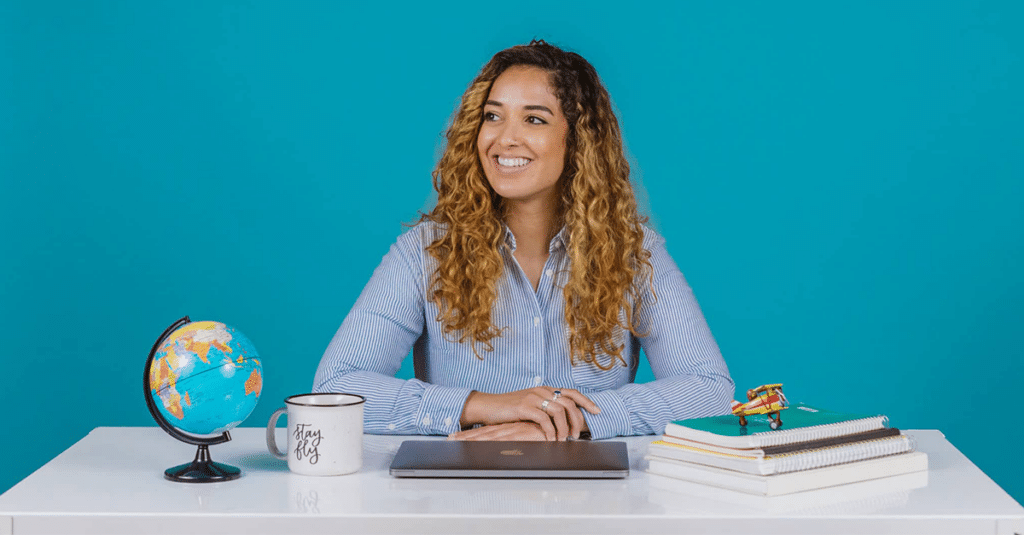 a woman with long curly hair sits at a desk in front of a blue background. A globe, a coffee mug, and a small stack of books sit on the desk