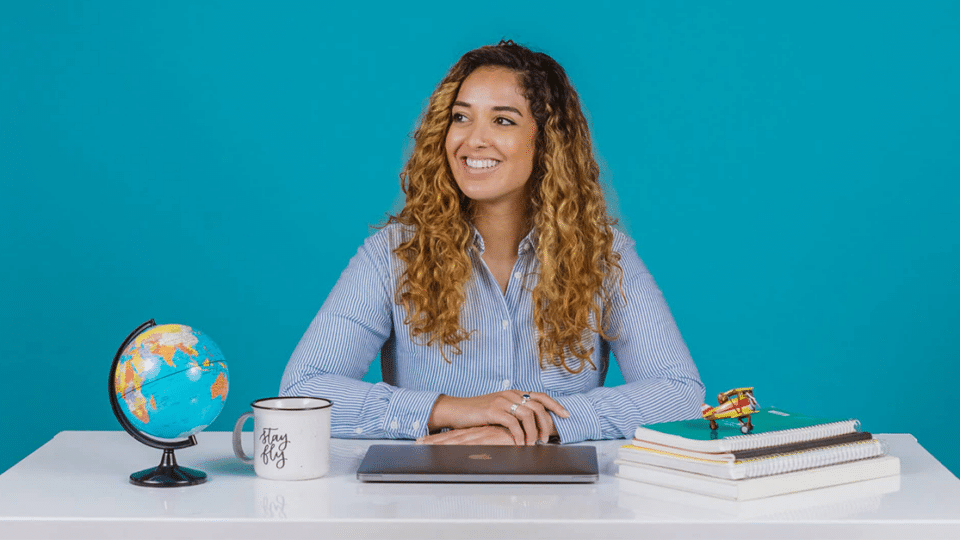 a woman with long curly hair sits at a desk in front of a blue background. A globe, a coffee mug, and a small stack of books sit on the desk