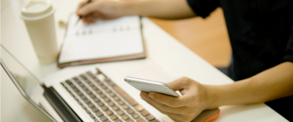 Photo of a person working on laptop and phone while writing in notebook