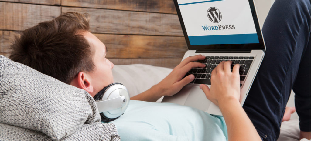 Photo of man lounging in bed using laptop to access WordPress with headphones