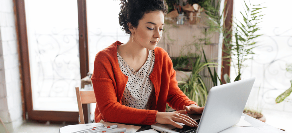 young woman with dark curly hair wearing a red cardigan works on a laptop at a cluttered work station