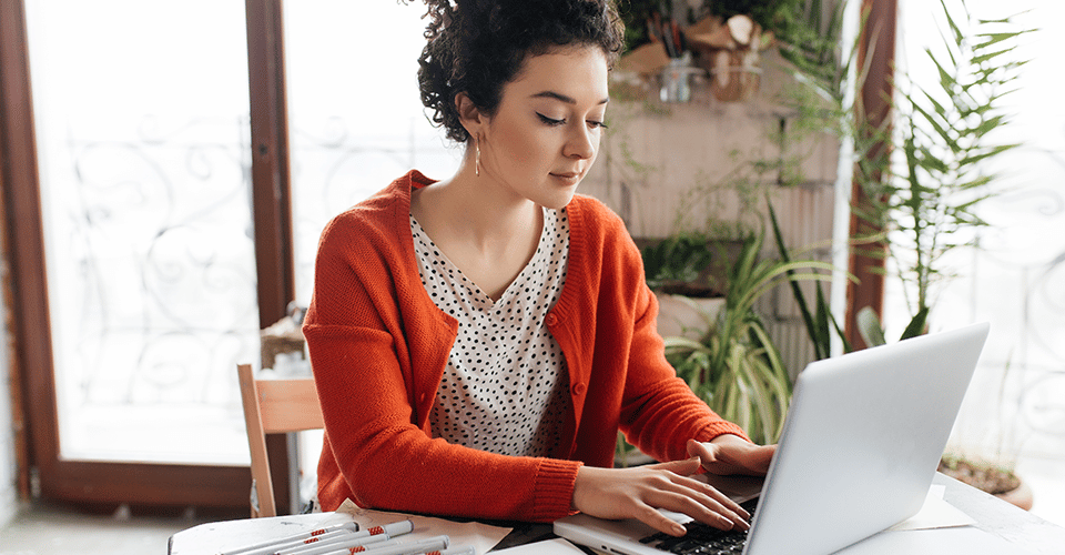 young woman with dark curly hair wearing a red cardigan works on a laptop at a cluttered work station