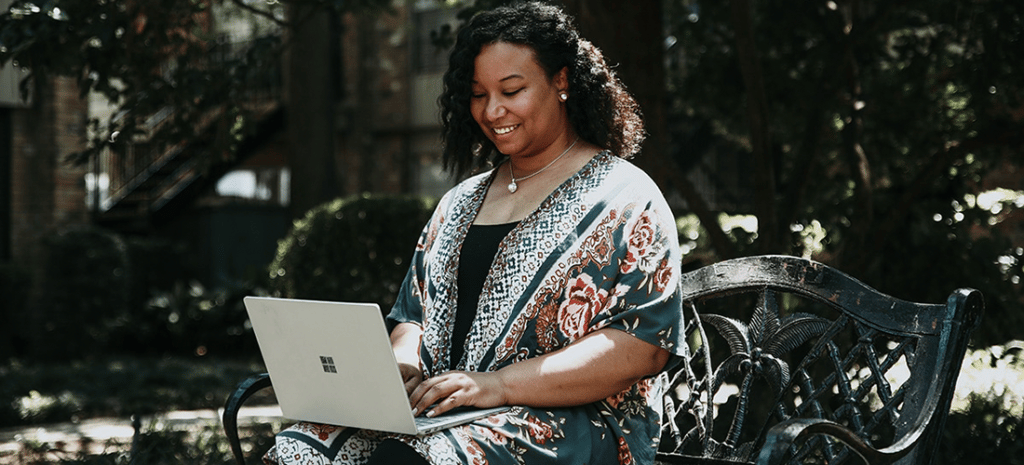 a Black woman sits on an outdoor bench on a sunny day while working at her laptop and smiling