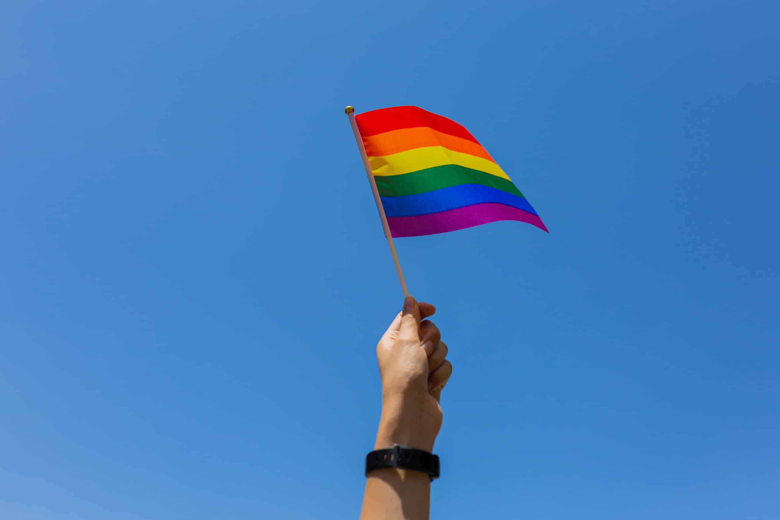 a hand holds a small Pride flag in front of a clear blue sky