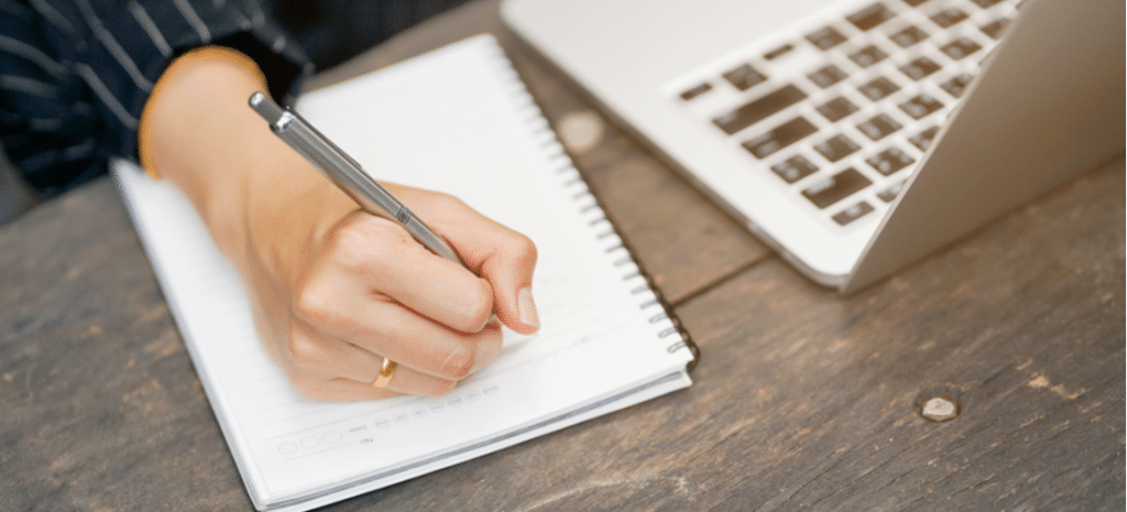 person writing in notebook next to macbook