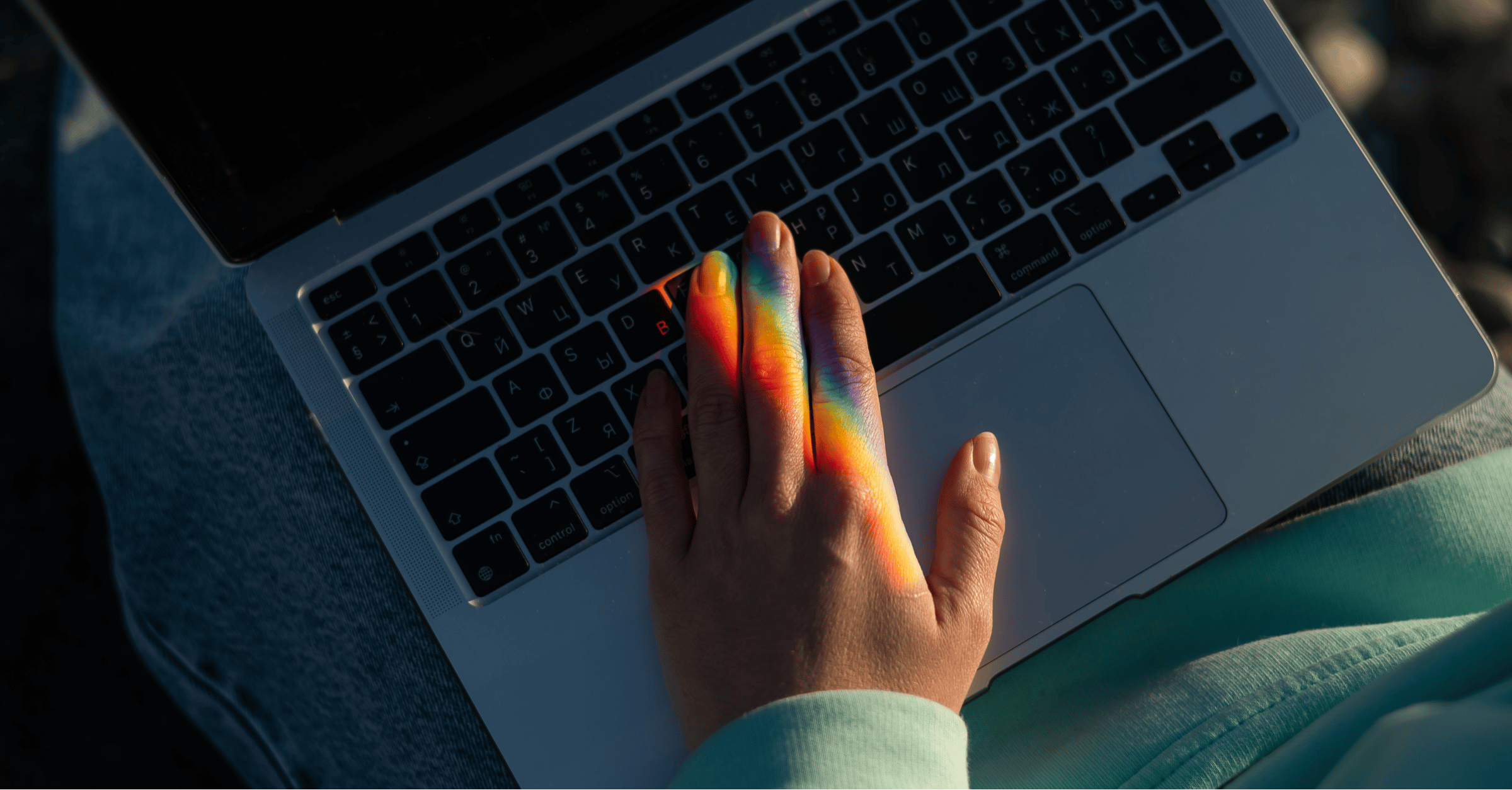 a person's hand hovers over a laptop keyboard. a rainbow prism falls over their fingertips
