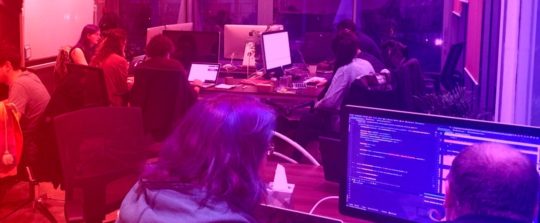 Participantes de um evento Queer Code trabalham em seus computadores em mesas de grupo