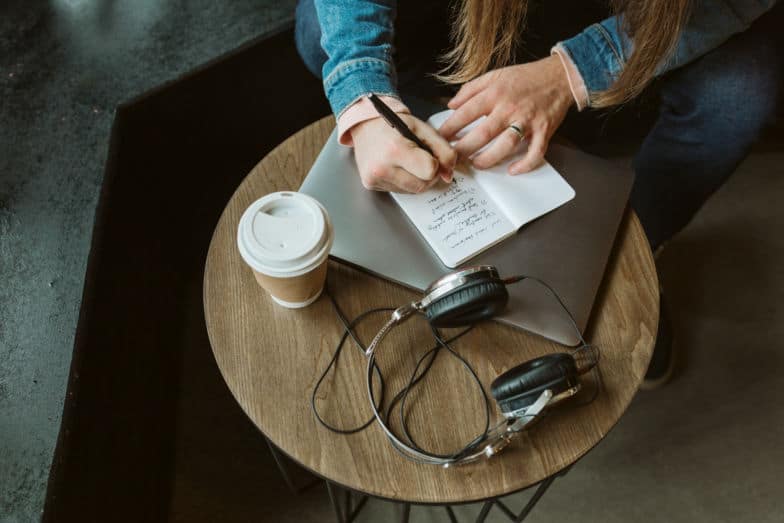 a person writes a quick note in a notebook which sits on top of a closed laptop next to a pair of headphones and a to-go cup of coffee