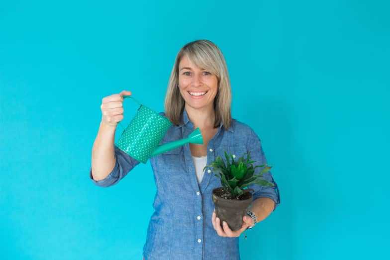 a woman holds a polka dotted watering can in her right hand and a potted plant in her left