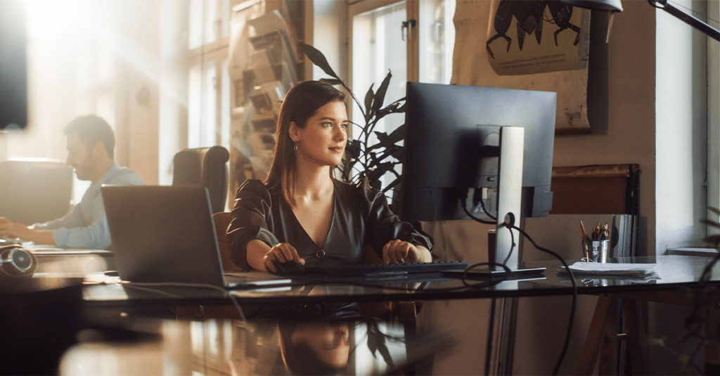 Woman working at computer in office setting, man working at computer in background